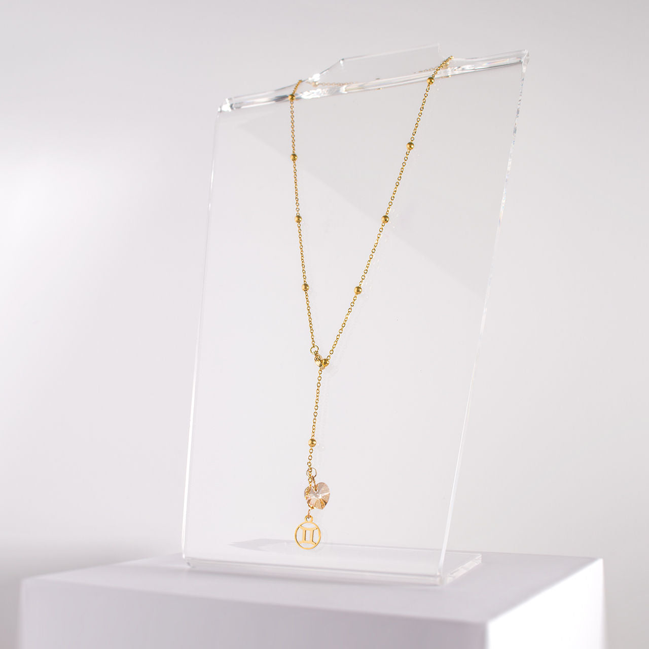 an 18K gold over stainless steel beaded station lariat necklace with a lobster claw clasp, Gemini charm and golden crystal heart on a clear acrylic stand