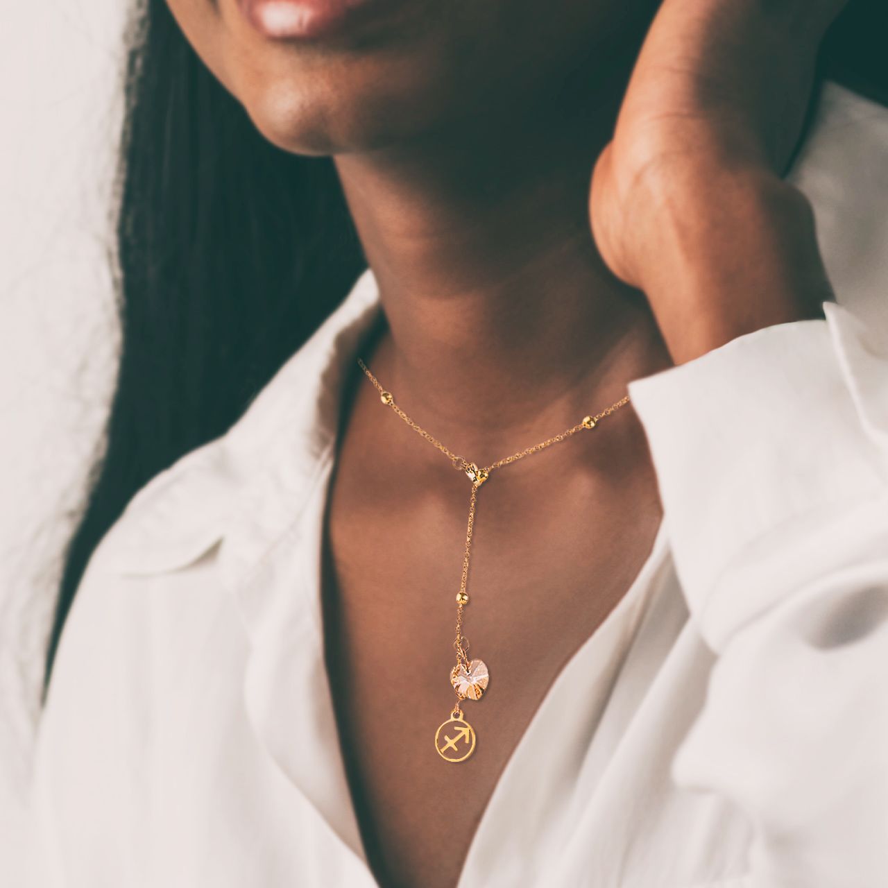 an 18K gold over stainless steel beaded station lariat necklace with a lobster claw clasp, Sagittarius charm and golden crystal heart on a dark skinned model neckline